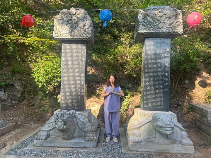 Visitor in temple stay clothing standing prayerfully between two tall stone tablets engraved with Hanja.