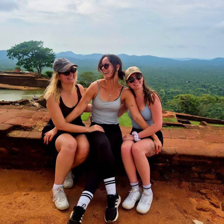 Three travellers relax and laugh on ancient red-brick ruins overlooking lush forest.