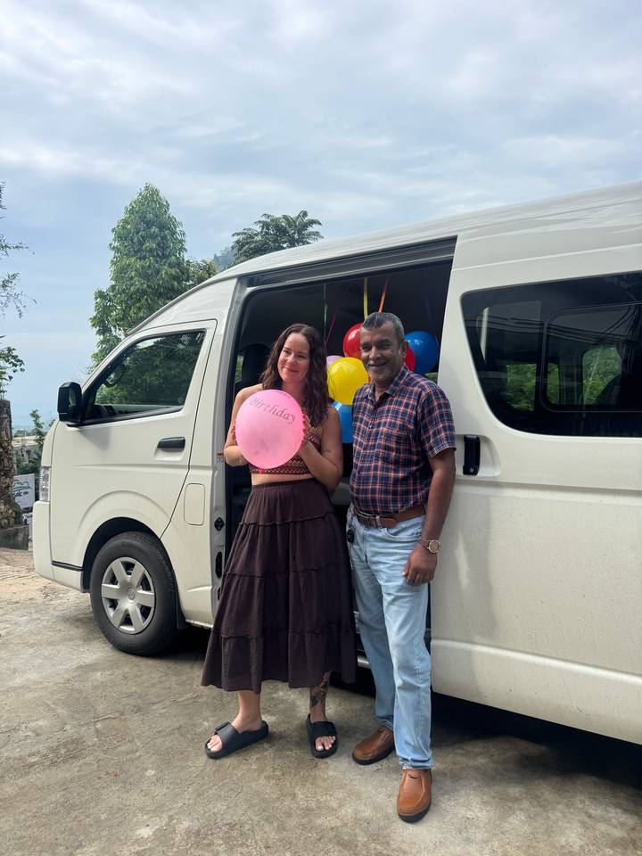 Traveller with birthday balloon poses beside a van decorated with colourful balloons.