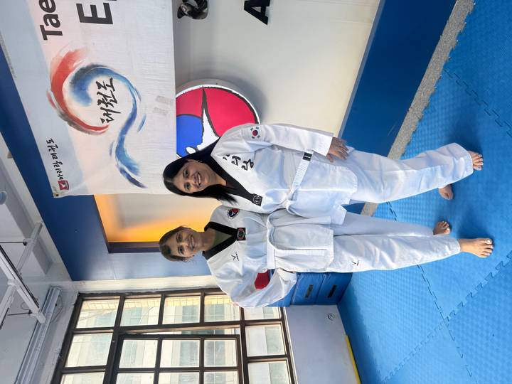 Two women wearing white taekwondo uniforms smile on a blue mat inside a martial arts studio.