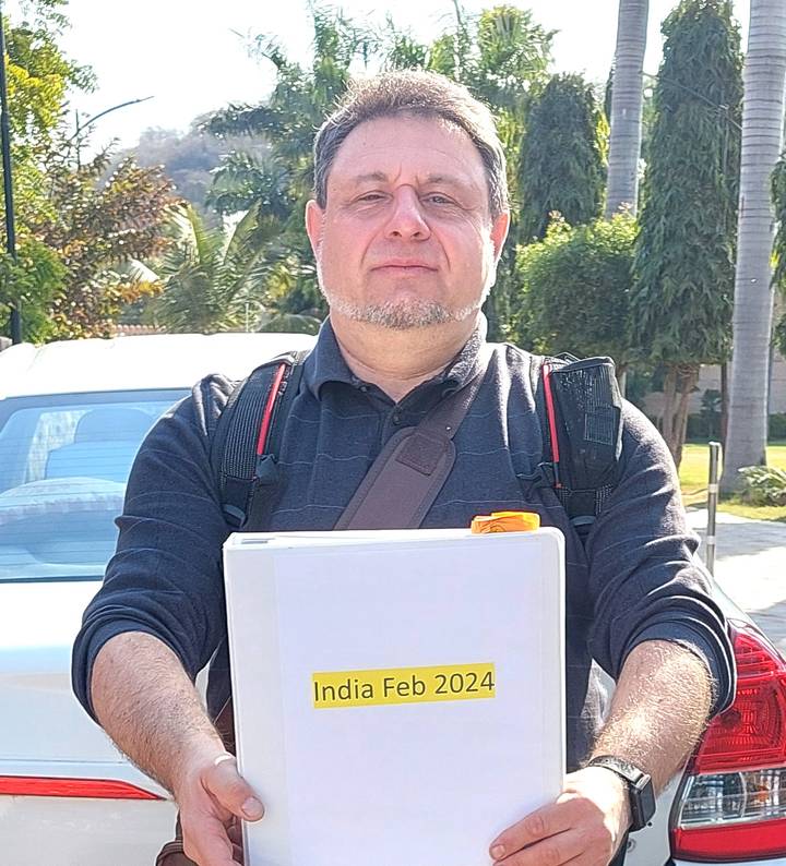 Close-up outdoor portrait of a man holding a folder, wearing backpack straps in bright sun.
