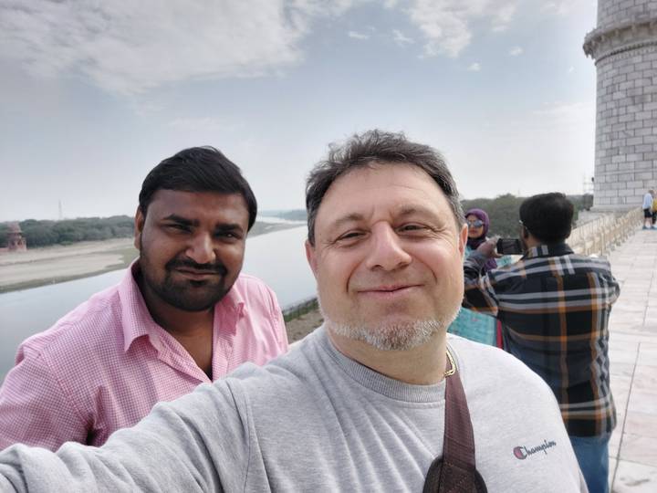 Casual outdoor selfie of two men smiling by a riverside monument with other tourists behind.