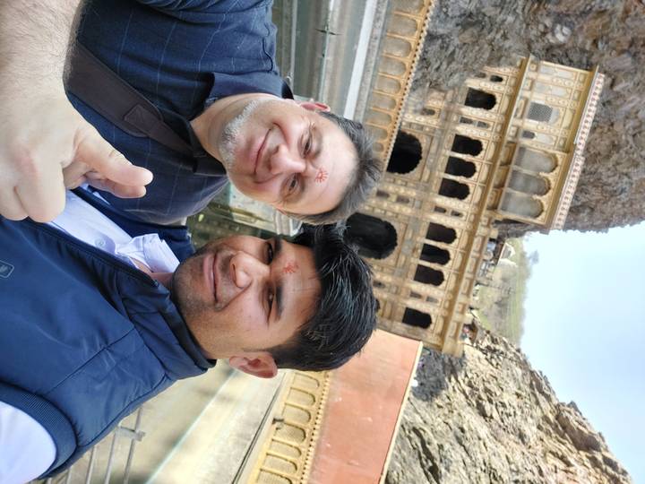 Two men with tilak marks posing for a selfie in front of ornate temple architecture