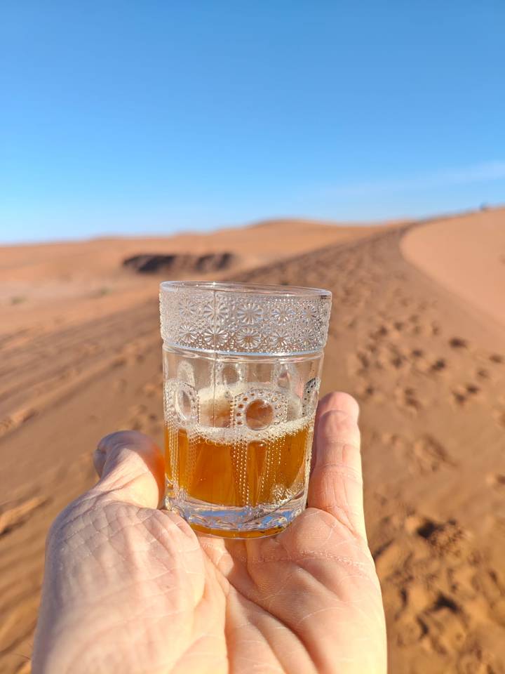 Hand holding a decorated glass of tea against sweeping Sahara sand dunes at golden light