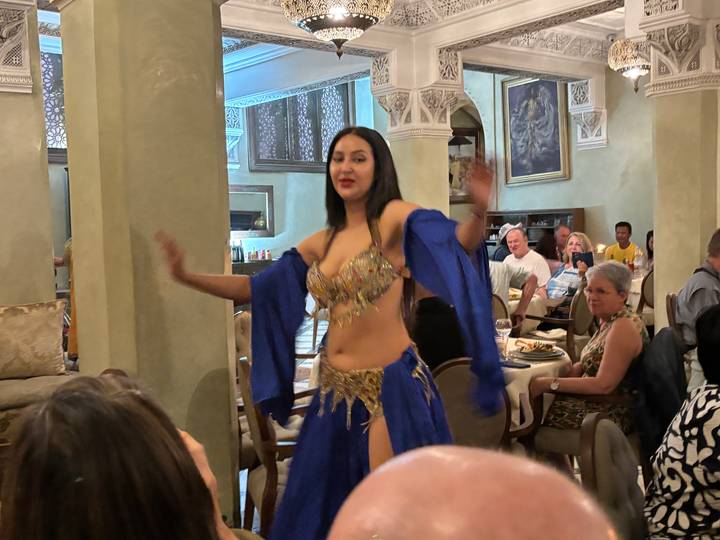 A belly dancer in a blue costume performs for diners inside an ornately decorated restaurant.