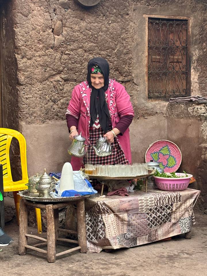 An elderly woman in a pink cardigan pours mint tea from two ornate silver teapots onto a tray of glasses in a rustic courtyard setting.