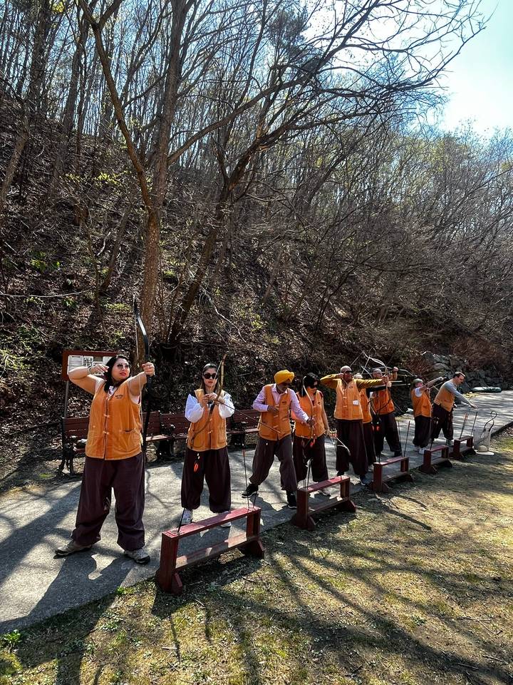A group of visitors wearing matching orange vests practice archery at an outdoor range backed by a wooded hillside.