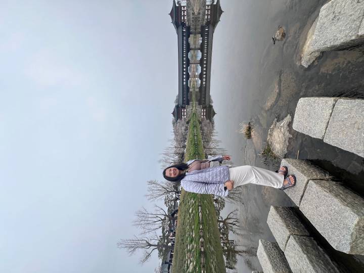 A woman poses on stepping stones beside a calm river with a traditional wooden bridge and stone walls in the background.
