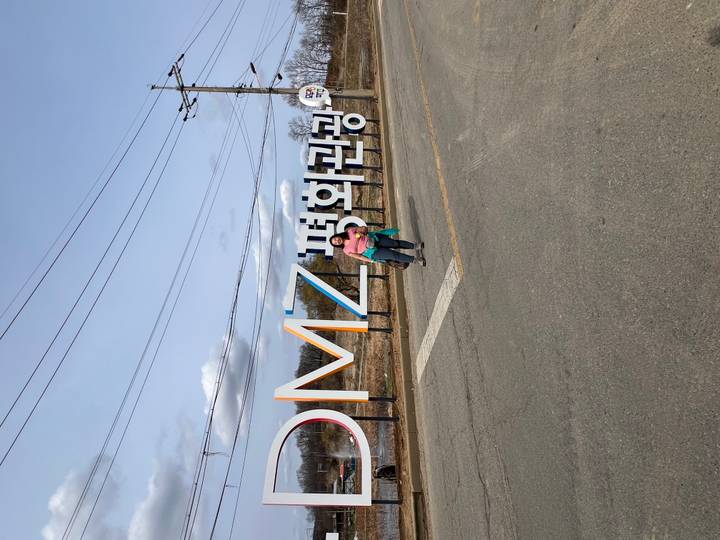 Two travellers stand in front of a large roadside DMZ peace tourism sign under a bright sky.