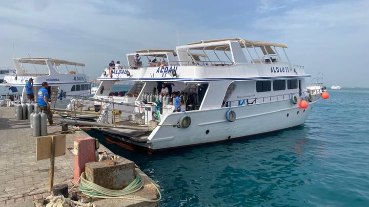Dive boat moored at a pier on clear turquoise water with scuba tanks and passengers preparing.