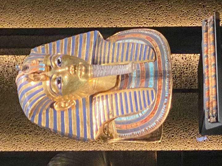 Golden funerary mask of Tutankhamun displayed behind glass in a museum setting.