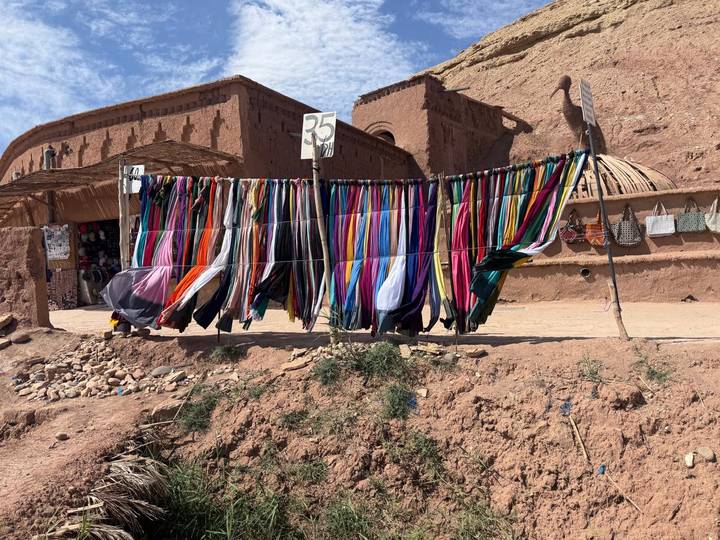 Rows of vibrant scarves hang for sale against the earthen walls of a desert village market.