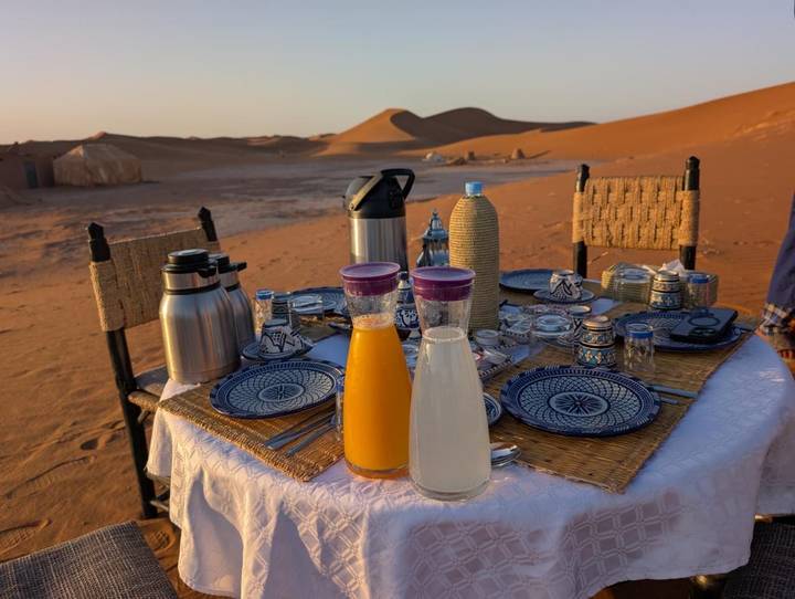 A neatly set breakfast table waits on desert sand with soft dunes and tents in the background.