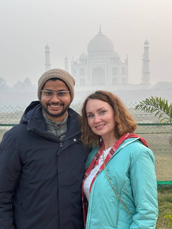 A smiling couple poses with the mist-shrouded silhouette of the Taj Mahal visible behind them.