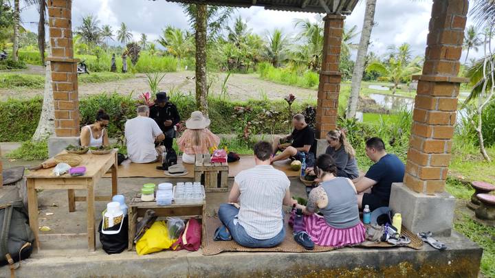 Travelers sitting on mats under open pavilion enjoying picnic in rice field setting.