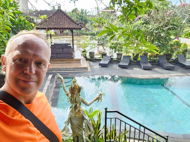 Selfie of traveler on balcony overlooking tranquil pool with sun loungers and lush greenery.