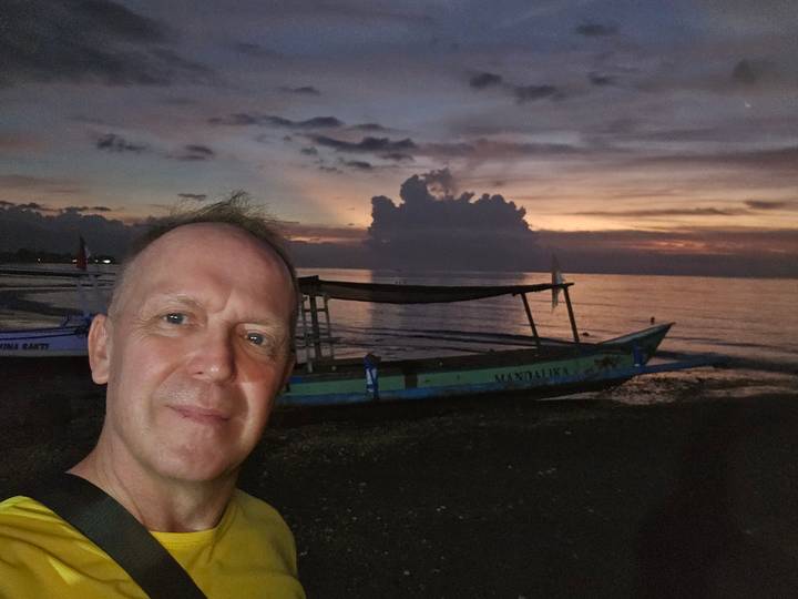 Low-light selfie on beach at sunrise with small fishing boat and colorful sky.