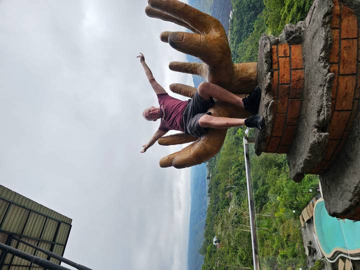 Man joyfully posing on giant hand sculpture viewpoint above lush valley.