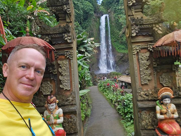 Selfie at ornate Balinese gate framing tall rainforest waterfall.
