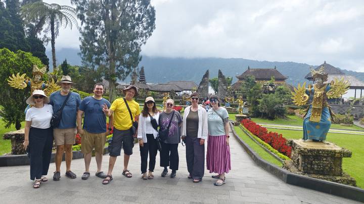 Group of travelers posing in front of ornate Balinese temple gardens with misty mountains behind.