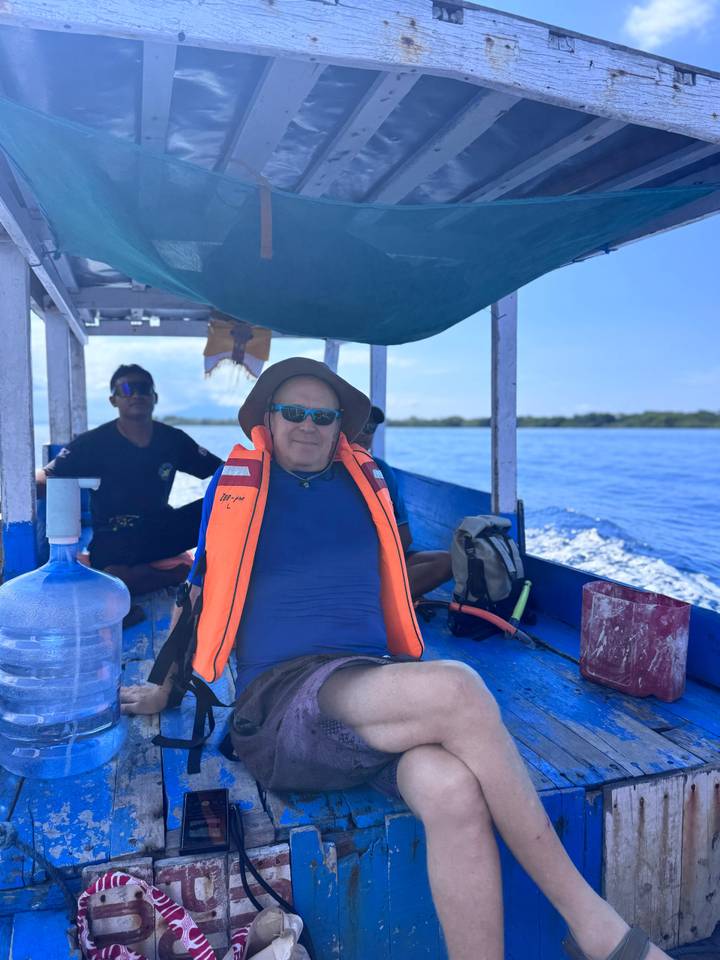 Man wearing life vest relaxing on blue wooden boat with captain behind on calm sea.