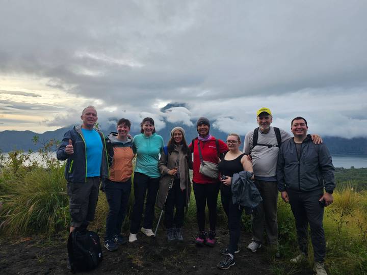 Group of trekkers posing on grassy ridge with dramatic cloud-covered volcanic lake backdrop.