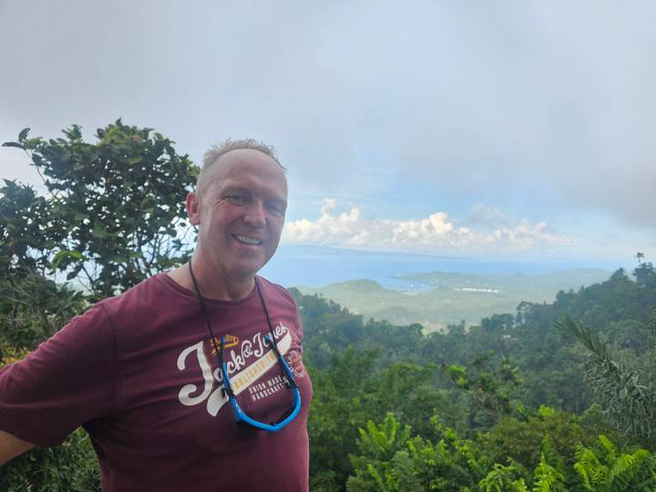 Smiling hiker with panoramic view of forested hills and sea in background.