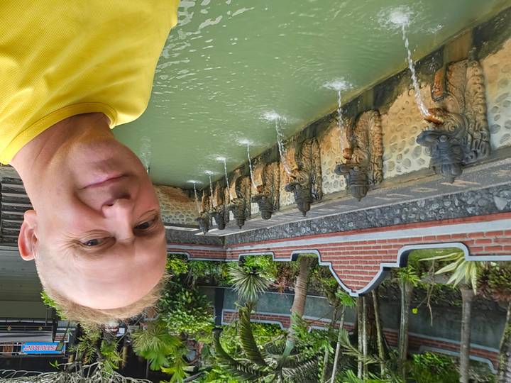 Man smiling beside a stone fountain lined with dragon-head spouts pouring water into a green pool at a Balinese temple.