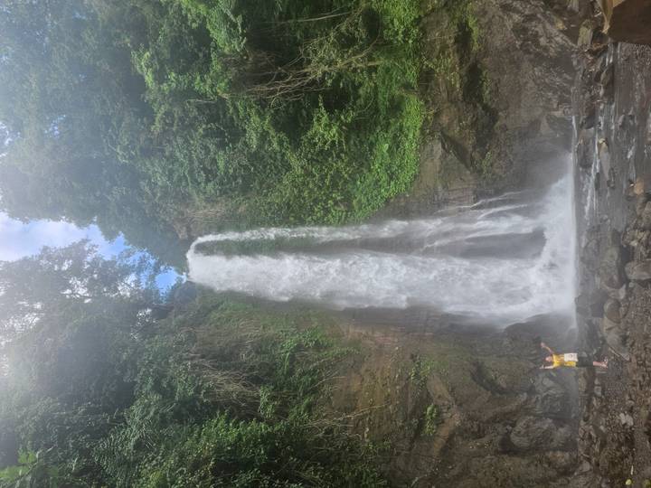 Tall waterfall plunging down a lush cliff into a rocky pool with a lone person in a yellow shirt posing at the base.