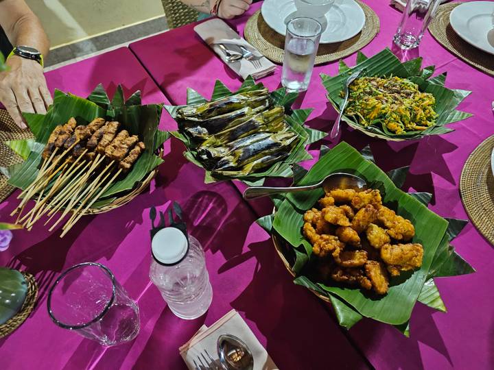 Assorted Indonesian dishes—satay skewers, grilled banana-leaf parcels, stir-fried greens, and fried bites—served on banana leaves over a magenta tablecloth.