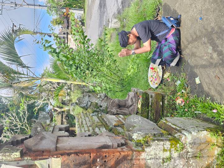 Balinese man in traditional attire kneeling in prayer before a small roadside shrine with fresh offerings under bright sunlight.