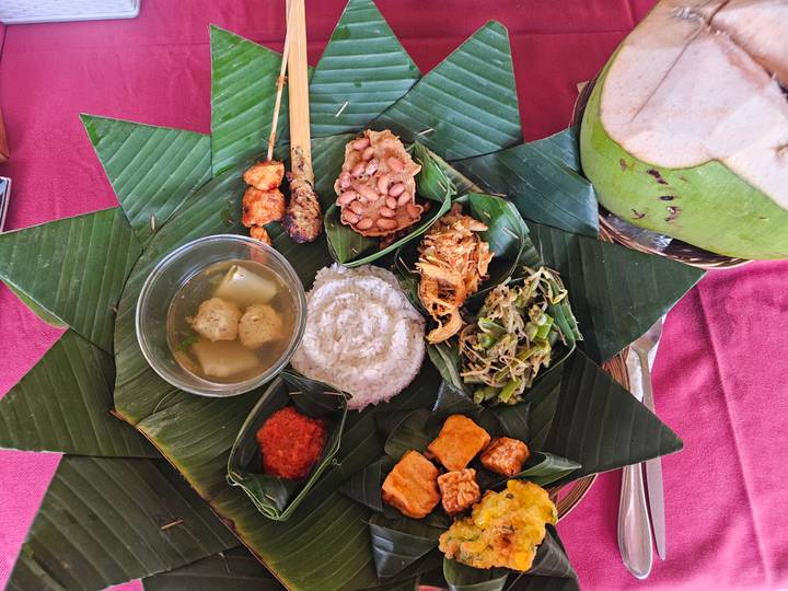Traditional Indonesian meal arranged on banana leaves with rice, soup, grilled skewers, vegetables, coconut and assorted side dishes.