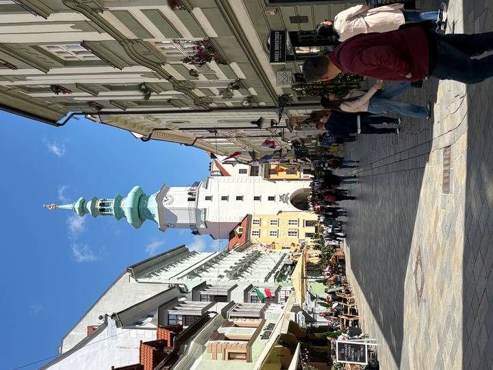 Lively pedestrian street lined with colorful historic buildings and café terraces leading to a green-roofed tower gate