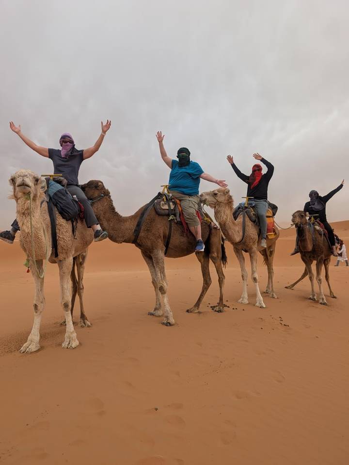 Five travelers riding camels raise their arms while traversing orange Sahara dunes.