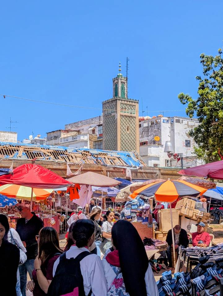 A vibrant street market with colorful umbrellas set below a tall green-tiled minaret amid whitewashed buildings.