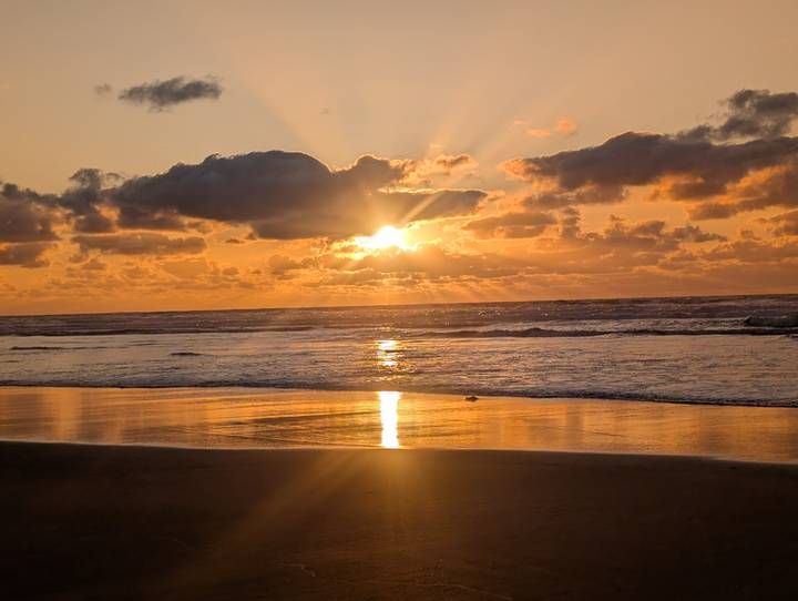 Golden sun sets over a tranquil beach, its reflection glowing on the wet sand beneath dramatic clouds.