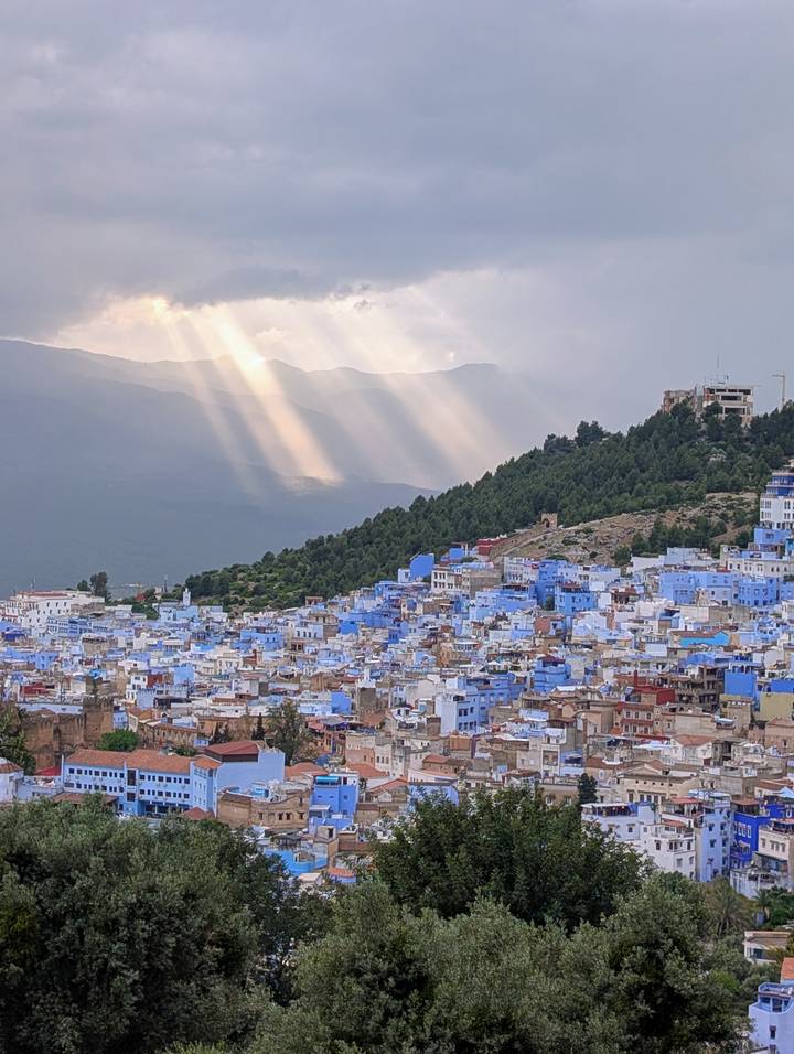 Chefchaouen’s blue-painted houses sprawl across a hillside as sunbeams pierce clouds above the Rif Mountains.