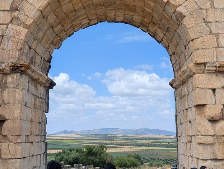 Stone Roman arch at the ruins of Volubilis frames rolling farmland and distant mountains under a blue sky.