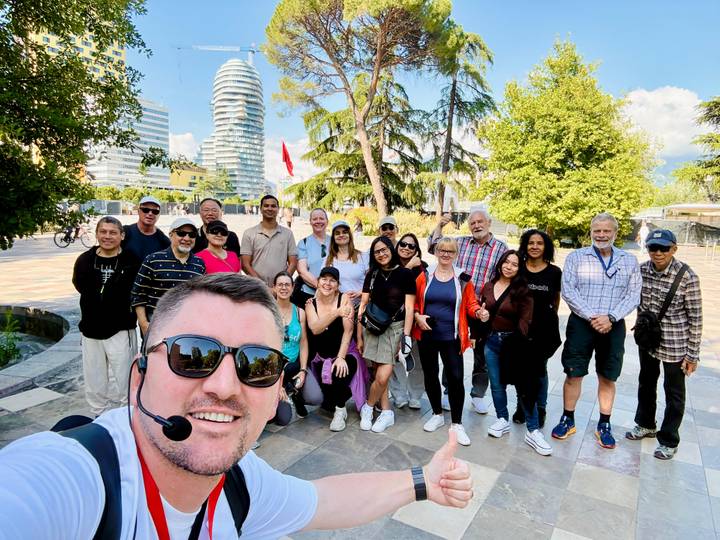 Cheerful selfie of a tour group in a modern city park with a distinctive curving skyscraper in the background.