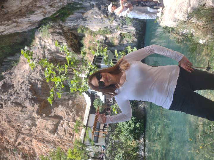 Woman posing beside a turquoise spring and cliffside Tekke house at Blagaj.
