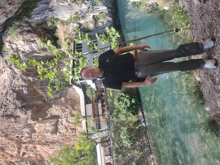 Man posing beside clear spring water and a cliffside restaurant similar to Blagaj’s setting.
