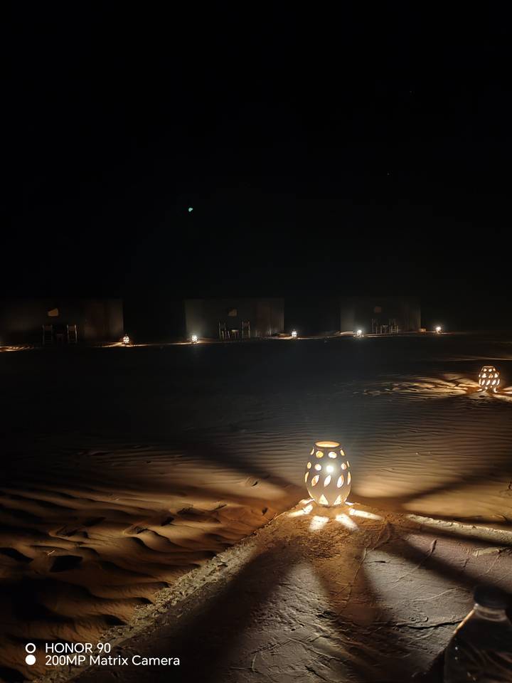 Nighttime desert camp with lanterns illuminating sand ripples and simple tents in darkness.