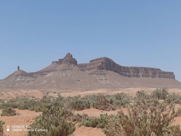Wide view of a rugged mesa-like rock formation rising from a barren desert plain under a bright blue sky with sparse shrubs in the foreground.