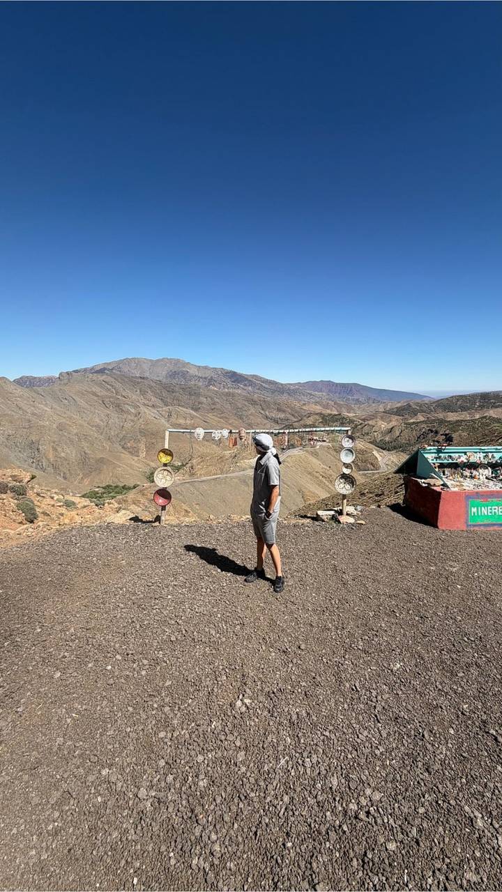 Traveler in headscarf surveys a winding mountain valley from a high roadside viewpoint lined with colorful souvenir plates.