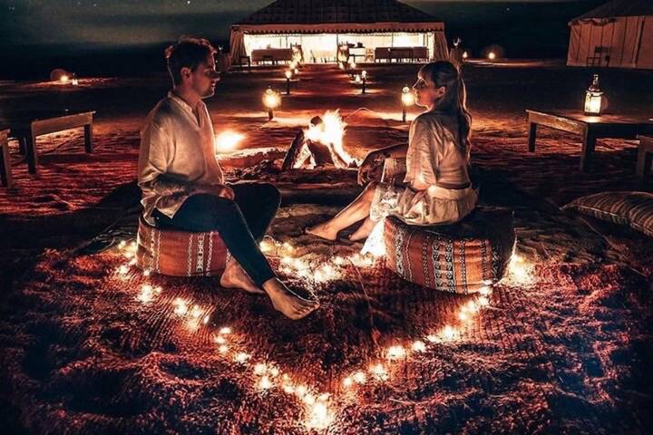 Couple seated on cushions inside a heart-shaped ring of fairy lights beside a campfire at a desert glamping site at night.