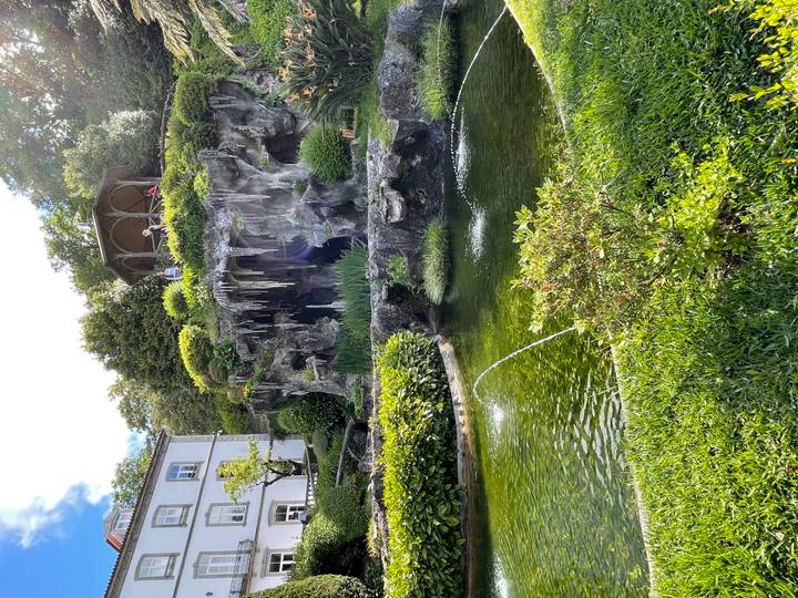 Lush garden with artificial grotto, pond and spraying fountain beside a stately home.