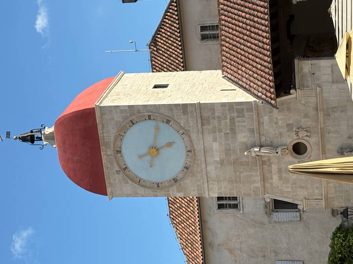 Stone clock tower with a red domed roof and pale blue clock face against clear sky.