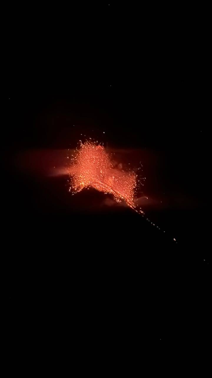 Night eruption with bright orange lava fountain streaming down volcano slope against black sky.