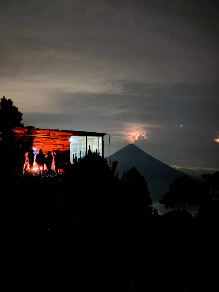 Silhouetted hikers warming by a fire outside a modern cabin overlooking a conical volcano glowing in the night sky