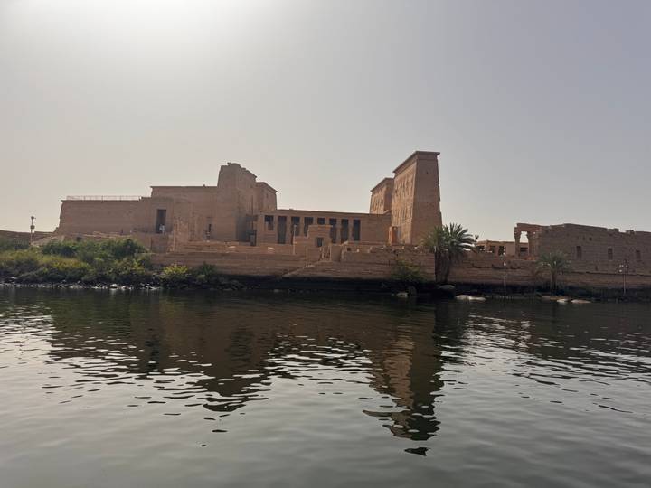 Ancient sandstone temple complex sitting on a small island, reflected in calm river water under a clear sky.
